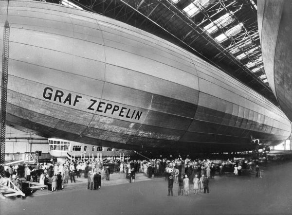 Slightly elevated view of a crowd surrounding the Graf Zeppelin inside a hangar. (It was named after the German pioneer of airships, Ferdinand von Zeppelin, who held the rank of Graf  or Count in the German nobility.) During its operating life the great airship made 590 flights, covering more than a million miles, including a round-the-world flight starting on 1929 at Lakehurst Naval Air Station, NJ.