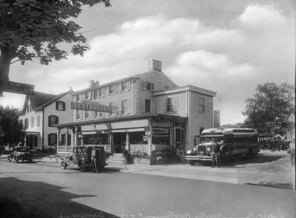View across street toward the Center Restaurant and Bus Terminal, with automobiles and a bus parked nearby. The bus driver is standing in front of the bus, which displays "New York" above its front window. A woman is seated next to the bus on a bench, and another man is posed standing in front of his automobile in front of the restaurant.