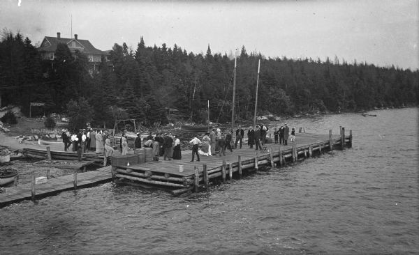 Elevated view toward a small boat landing and pier area on the shore of Lake Michigan. Many men, women and children are on the pier, and a house is on a hill behind trees in the background.