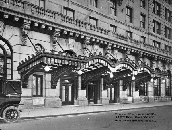 View from street toward the main entrance to the DuPont Hotel, a twelve-story Italian Renaissance hotel commissioned by DuPont Company president Pierre S. du Pont. In the foreground on the left is an automobile parked along the curb. Caption reads: "Main Entrance, Hotel DuPont, Wilmington, Del."