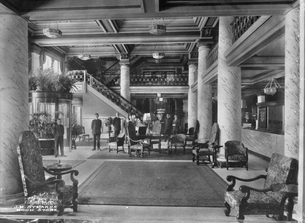 An interior view of a hotel lobby, filled with chairs and other furniture, plants, columns, and several men standing in the background.