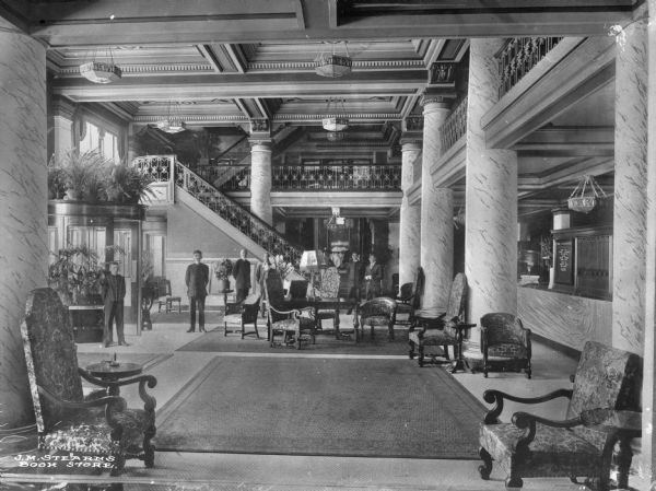 A view of a hotel lobby filled with chairs and other furniture, plants, columns, and several men standing in the background.