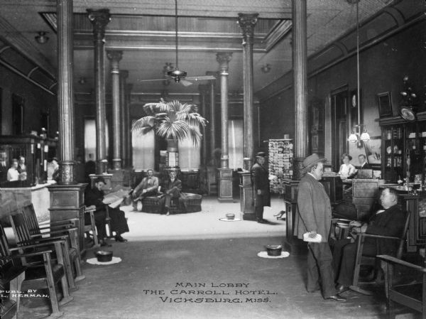 The main lobby in the Carroll Hotel, named for Capt. E.C. Carroll. People are sitting and standing in the lobby which has tall ceilings, columns, and chairs. Two people are standing behind the desk on the right, and two more people are standing behind another desk on the left. The Carroll Hotel was razed in the 1960s to make space for a parking garage. Caption reads: "Main Lobby, The Carroll Hotel, Vicksburg, Miss."