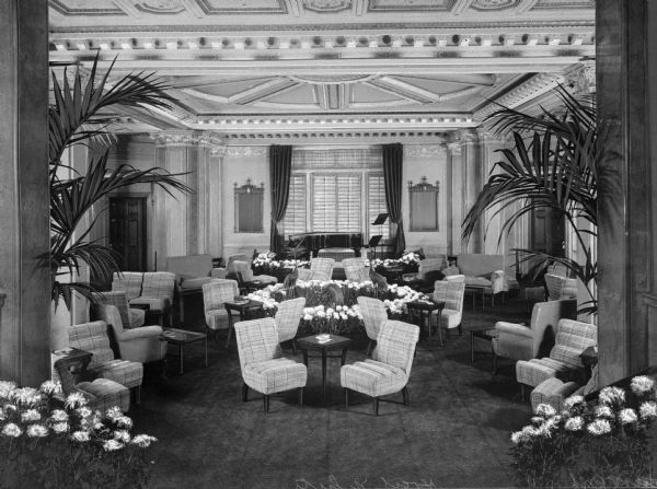 Slightly elevated view of the lobby of the Hotel White. Tables, chairs and potted flowers and plants are arranged in the foreground, and in the background a piano is centered in front of a window framed with drapes.