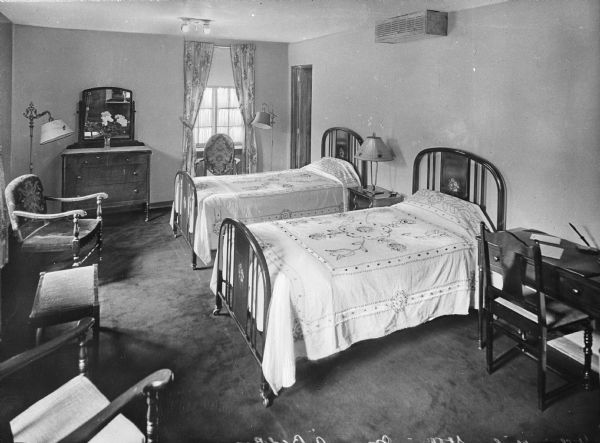 A view of a bedroom at the St. Clair Inn. It features two beds, a dresser, mirror, a nightstand, lamps, a desk, and chairs.