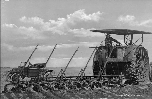 A view of two men on a steam tractor with a gang plow in a field. An automobile is behind the plow.