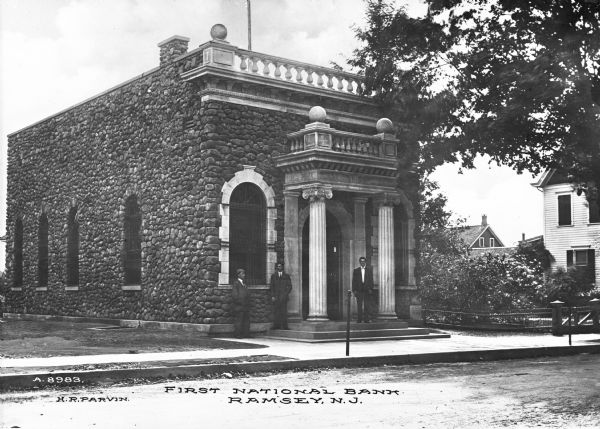 View from street toward three men standing outside of the First National Bank, a square, stone building with cobblestone construction. Caption reads: "First National Bank, Ramsey, N.J."