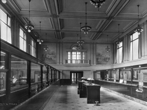 View of the interior of the First National Bank. Teller windows are on both sides of the room which has high ceilings.