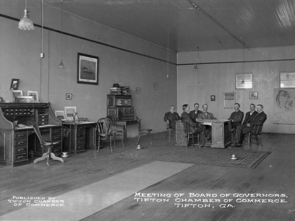 View across room toward a group of men sitting around a desk for a meeting. Caption reads: "Meeting of Board of Governors, Tifton Chamber of Commerce, Tifton, GA."