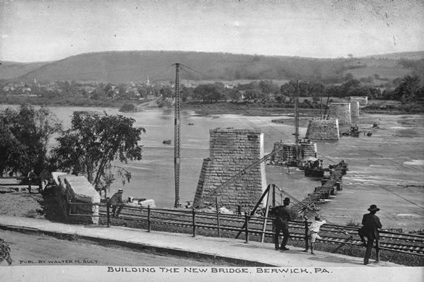 Slightly elevated view of several people overlooking a bridge under construction. Caption reads: "Building The New Bridge, Berwick, PA."