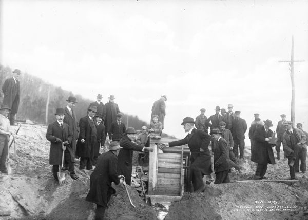 A view of a group of men wearing long overcoats and hats, standing on rough ground. Two men in the center are shaking hands, and a few others are holding shovels. They are possibly at the ceremony for the joining of the cuts for the Cape Cod Canal.