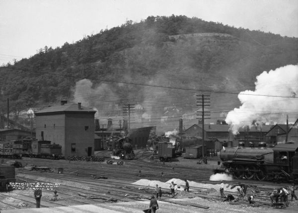 Elevated view of men working on the railroad tracks. There are locomotives and railroad cars on the tracks. Some children are standing on the tracks near the workers. In the background is a hill.