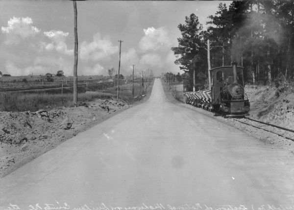 View down center of a rural section of U.S. highway, with a small train running on railroad tracks alongside it.