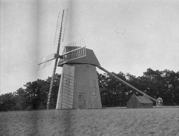 A windmill in an open field, with a pitched tent and trees in the background.