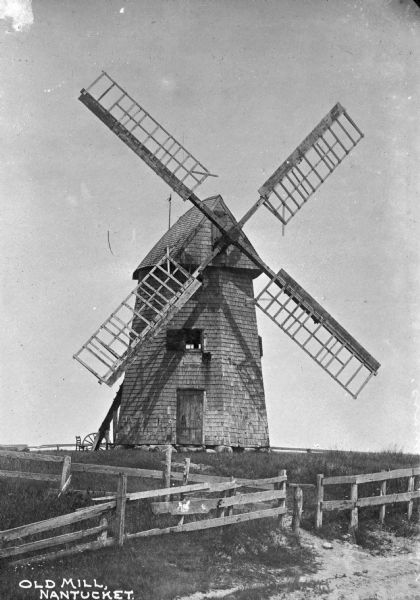 A view of an old windmill surrounded by wooden fencing in Nantucket, possibly Cottage City. Caption reads: "Old Mill, Nantucket."
