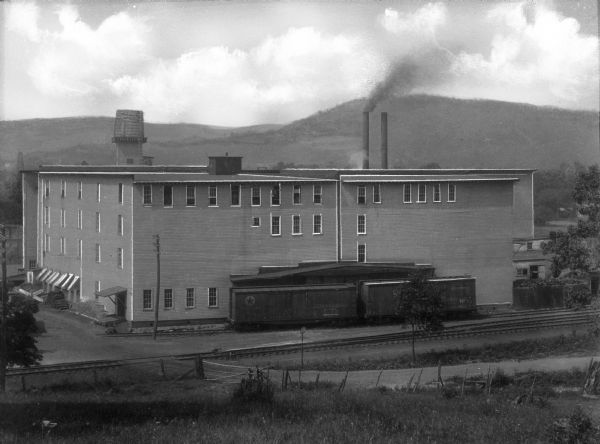 Elevated view of a factory in Youngsville, with a railroad car and railroad tracks in front of it. In the background is a hill or mountain.