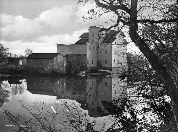 View across water toward an old factory. The factory buildings are reflected in the water.