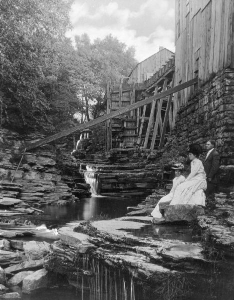 A portrait of two women and one man who are sitting on rocks near the waterfall. There is an old mill above them on the right.