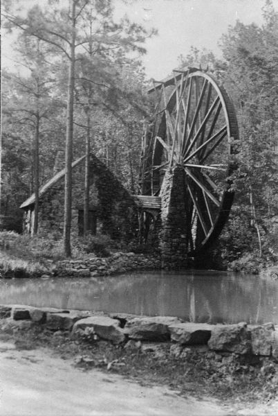 View from road with a stone wall toward a small stone building housing a grist mill, with a large water wheel on the right side. Surrounding the grist mill are trees and a small pond.