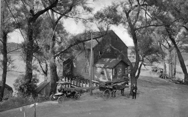 Elevated view of the old mill at State Mills Lake. A man, an automobile, and a horse and carriage are on the road in front of the mill.
