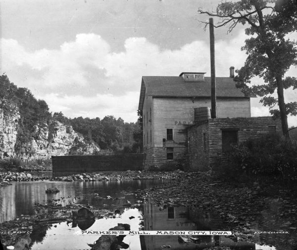 View across water with rocky shoreline toward the Parkers Mill. There is a rocky cliff in the background on the left. Caption reads: "Parker's Mill, Mason City, Iowa." 