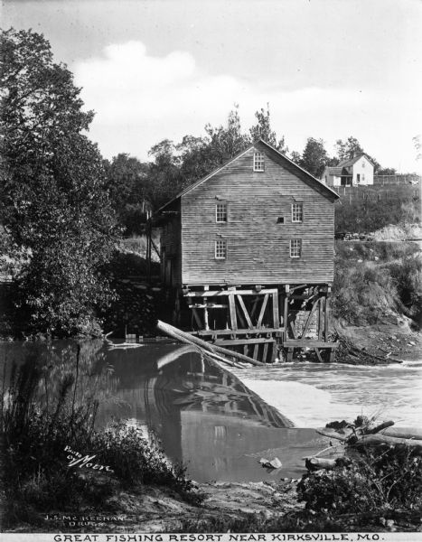 Elevated view of the banks along a fishing stream, with a few buildings on the other side of the water. Caption reads: "Great Fishing Resort Near Kirksville, MO."