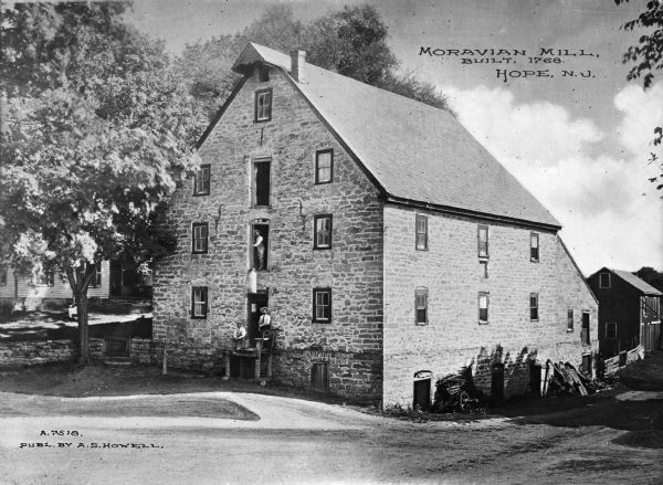 View across road toward a Moravian Mill, a large stone building. Two men are posed in the open door of an elevated landing of what may be a small loading dock. Another man is standing in the open doorway above them holding onto a rope attached to the roof overhang that is suspending a large sack just above the loading dock. A house is next to the mill on the left. Caption reads: "Moravian Mill, Built 1768. Hope, N.J."