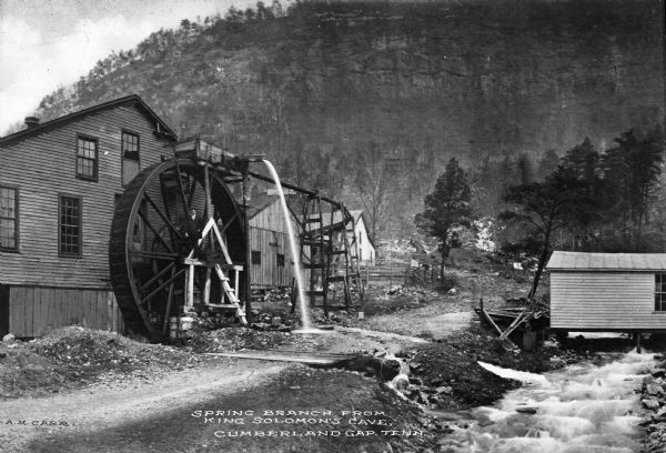 View toward a mill with a large water wheel and spring branch, near King Solomon's Cave. A man is posed standing next to the middle of the water wheel. Caption reads: "Spring Branch from King Solomon's Cave, Cumberland Gap, Tenn."
