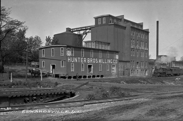 View across railroad tracks toward the Hunter Bros. Milling Co. mill. There is a railroad train on the tracks next to the mill. A man is standing in front of the mill and looking at two horses or possibly mules, that are pulling wagons ready to be loaded. Caption reads: "Edwardsville, Ill."