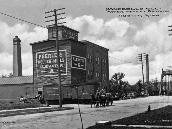A view of Campbell's Mill and the Water Street Bridge in the background on the right. There is a railroad car and several men driving horse-drawn wagons on the street. Caption reads: "Campbell's Mill, Water Street Bridge, Austin, Minn."