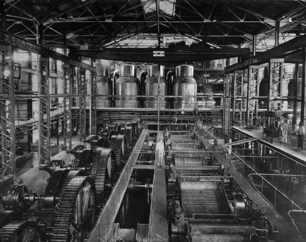 The interior of a sugar mill in Havana, Cuba, with a few men standing on scaffolding between the machinery.