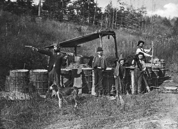 Two men,  three boys, and a dog, standing around an illegal moonshine liquor distillery in the mountains. The man posed on the right is stirring something in a barrel, and the man posed on the left is brandishing a gun.