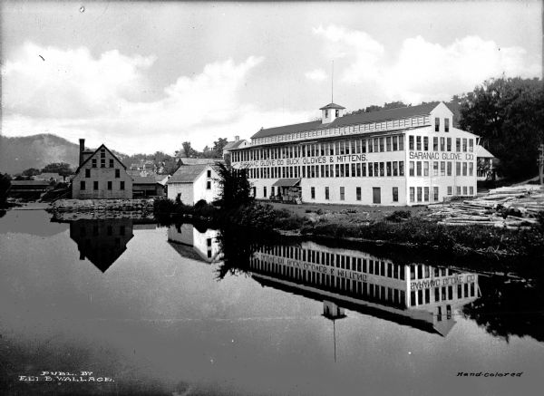A view of the Saranac Glove Co. factory, located next to a body of water, in which the factory's reflection is visible.