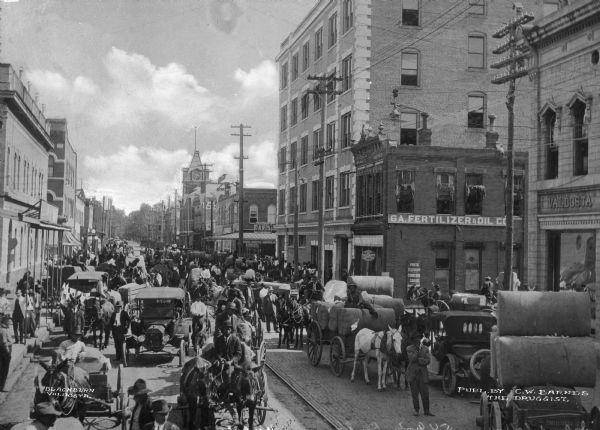 View of a street crowded with people, automobiles, horses and wagons. Many of the wagons are loaded with cotton. Text on the photograph reads, "Publ. by C.W. Barnes, the Druggist."