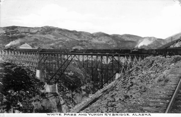 A view of a train crossing the Yukon Railway Bridge at White Pass, with pedestrians on one the side of the tracks. Caption reads: "White Pass And Yukon R.Y. Bridge, Alaska."