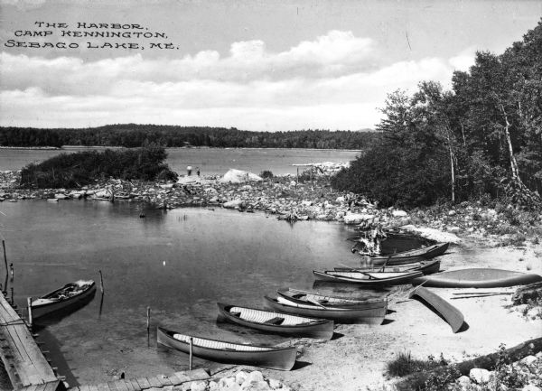 Elevated view of the harbor at Camp Kennington with boats pulled onto the shore of a small inlet, part of Lake Sebago. Caption reads: "The Harbor, Camp Kennington, Sebago Lake, ME."