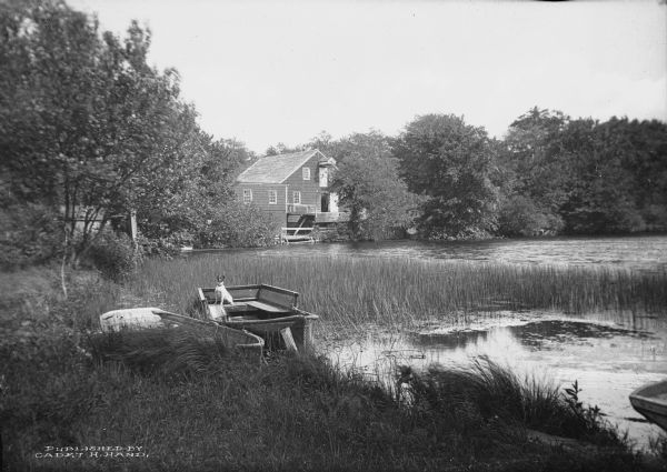 A peaceful view of a pond and mill, featuring a small dog sitting in a boat, which rests on the shore.