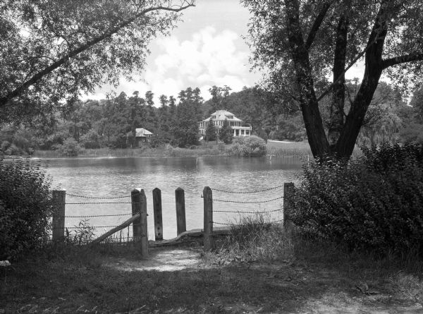 View across Scudder's Pond, featuring a fence framed by trees in the foreground, and a house on the opposite shoreline in the background.