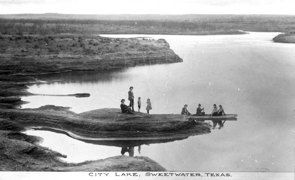 Elevated view of people sitting in a boat and and also standing on the shore of City Lake, with a view of the vast landscape in the distance. Caption reads: "City Lake, Sweetwater, Texas."