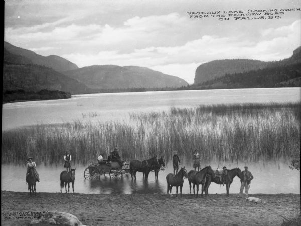 Children standing on horses' backs posing for the photograph, alongside of a wagon, and other horseback riders, on the shore of Vaseaux Lake. Caption reads: "Vaseaux Lake, looking south from the Fairview Road on Falls, B.C."