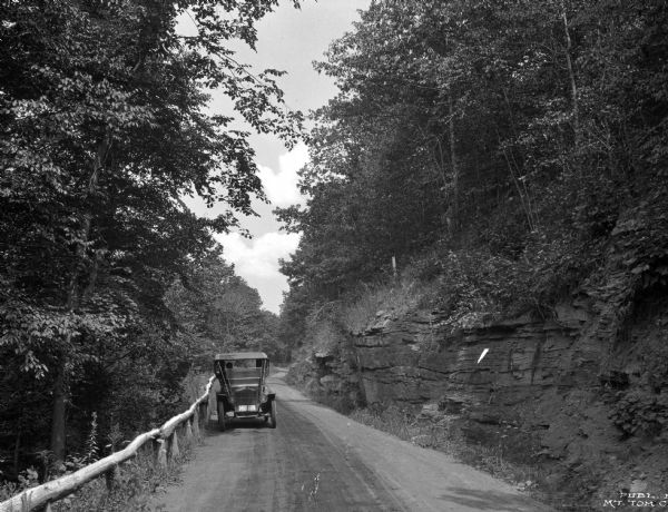 A view of a car on a dirt road driving toward Mt. Tom Camp, with a wooden guardrail on one side and a natural rock formation on the other side.