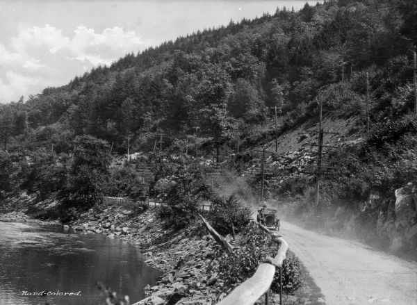 A view of a car driving on a dirt road along the Naugatuck River, with a wooden guardrail and river on one side, and a rock formation on the other.