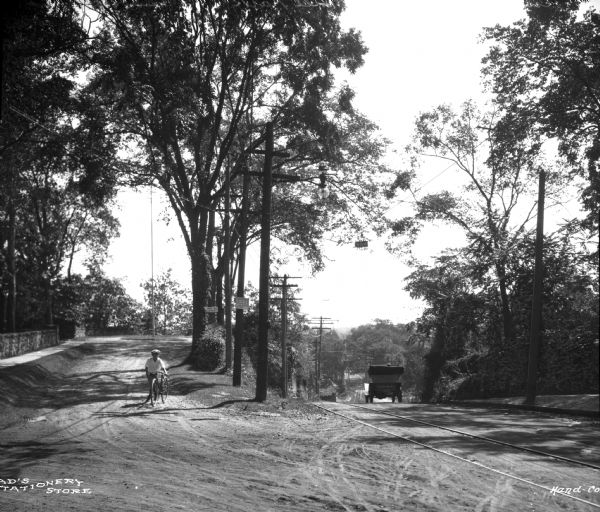 View toward a boy with his bicycle at the entrance to Putnam Hill Park on the left. There are telephone poles and trees in the center, street railroad tracks, and a car driving away on another road on the right.