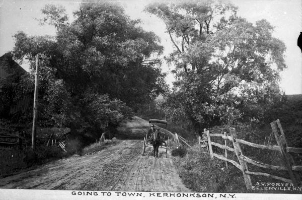 A view of a dirt road with a horse-drawn buggy, bordered by a split-rail fence, and trees. Caption reads: "Going To Town, Kerhonkson, N.Y."