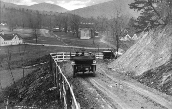 Elevated view of two cars on a dirt road heading downhill. A town is in the valley below, and mountains are in the distance.