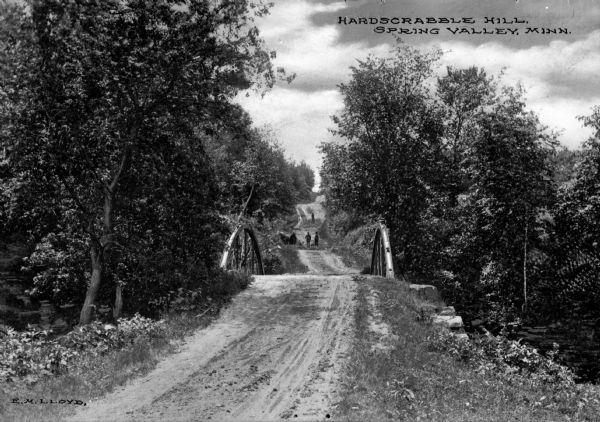 A view down a country dirt road near Hardscrabble Hill, which includes a small bridge and some people in the distance. The road is surrounded by trees on both sides. Text on photograph reads: "E.M. Lloyd." and "Hardscrabble Hill, Spring Valley, Minn."