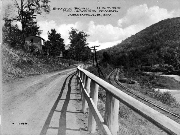 State road running alongside the Ulster & Delaware Railroad and the Delaware River. The dirt road is bordered by a fence on one side, and a hill with a house on the other side. Caption reads: "State Road, U. & D. R. R. Delaware River, Arkville, N.Y."