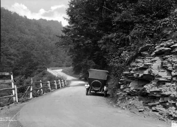 A view of a country dirt road with a car pulled over to the right side. The road is part of the Pennsylvania State Highway, and is bordered by a fence on one side, and rocks and trees on the other side.