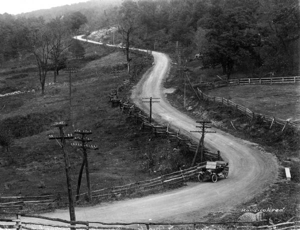 Elevated view of National Pike, on the west side of Martin's Mountain. The pike is a winding dirt road proceeding up a hill, surrounded by a fence and telephone poles on both sides.