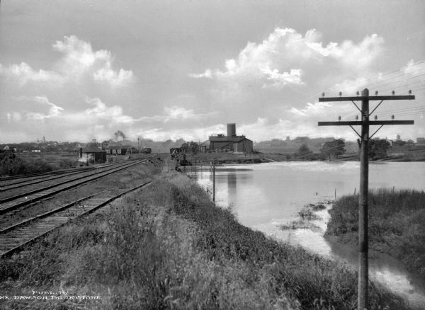 View of Rock Island Pond, with a telephone pole in the foreground, and railroad tracks leading off into the distance. In the distance is a farm or manufacturing building. 